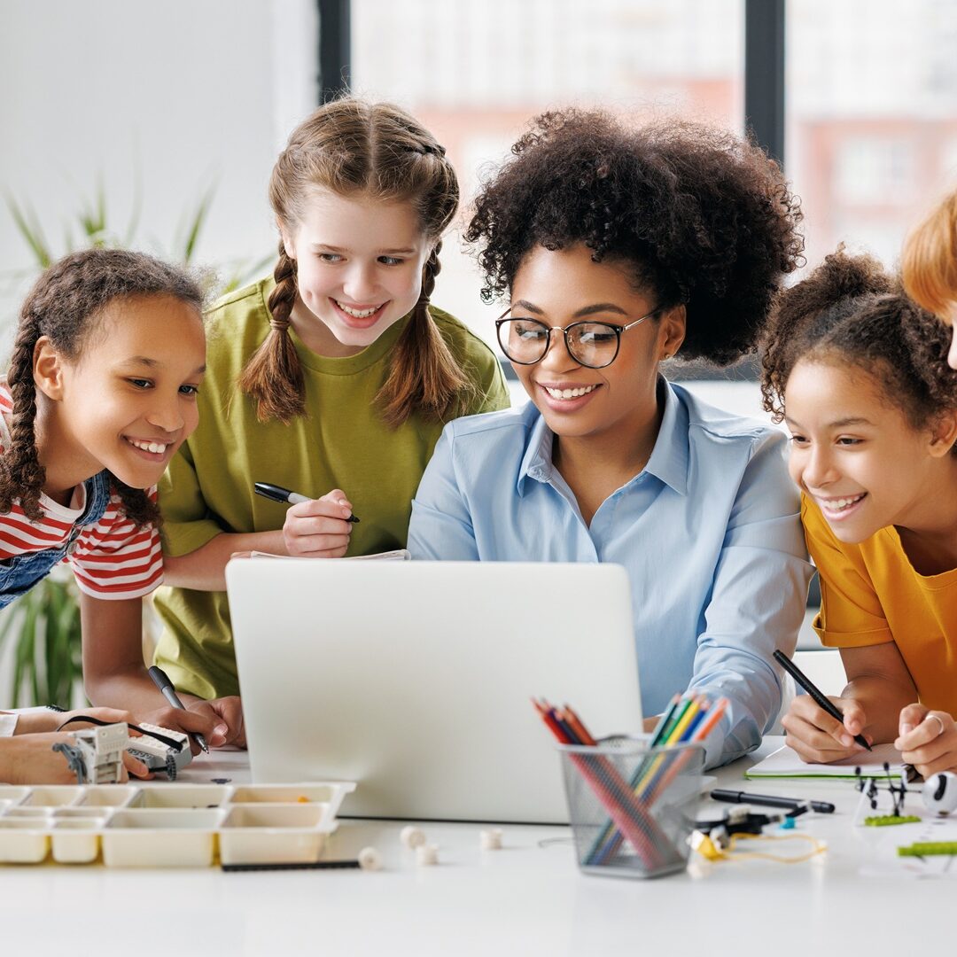 Happy female tutor and optimistic children smiling and watching laptop screen while gathering around table during lesson at school